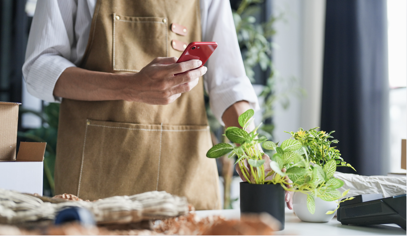 man in brown apron holding red phone taking photo of a plant on the counter in front of him